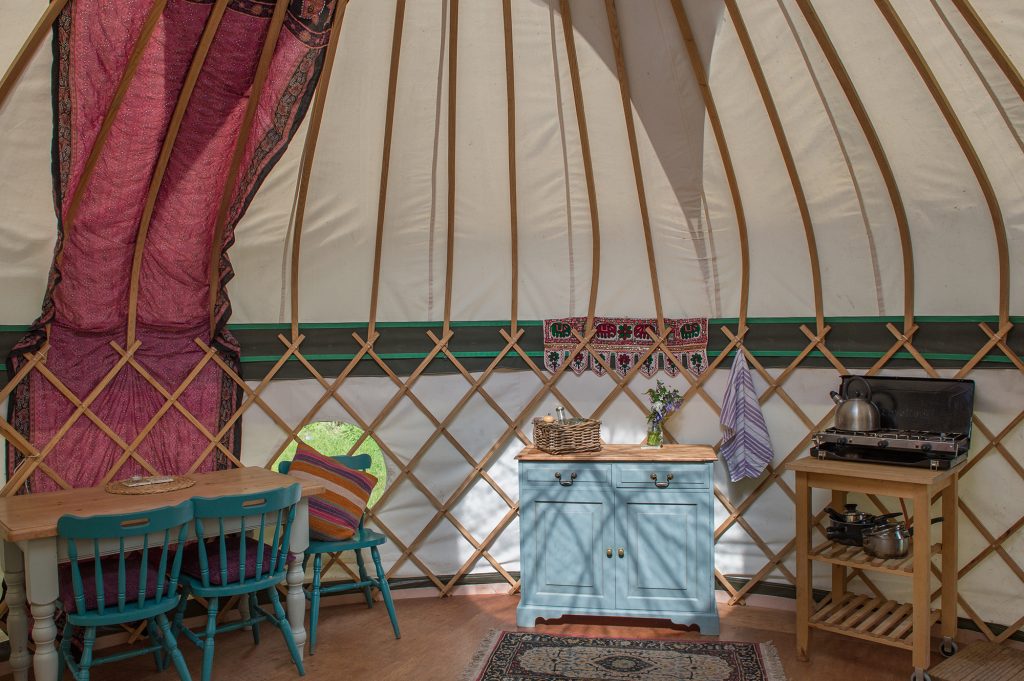 Luxury Fern Pippin Yurt interior with dining table, stove and cooking equipment, green sideboard with organic hamper and wild flowers at Cotna Eco Retreat near Mevagissey, Cornwall