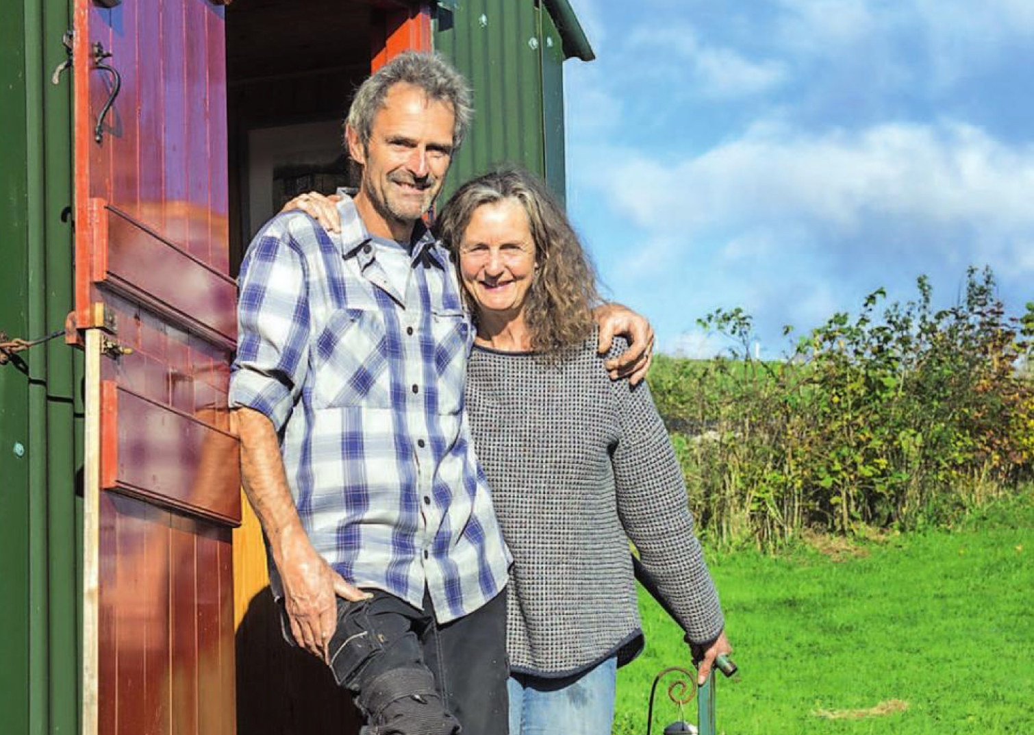 Owners Sara and Dave standing on steps of Ismay Shepherd’s Hut at Cotna Eco Retreat with green fields and blue sky behind them near Gorran Haven, Cornwall