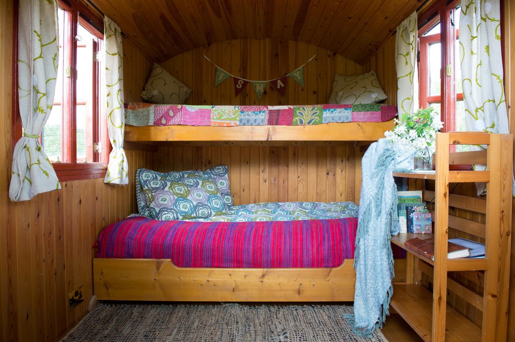Interior of Ismay shepherd’s hut at Cotna Eco Retreat on The Roseland, Cornwall, with organic seasalt sheets, pink Indian blanket on double bed and patchwork throw on single bunk bed. Wild flowers and home-made curtains